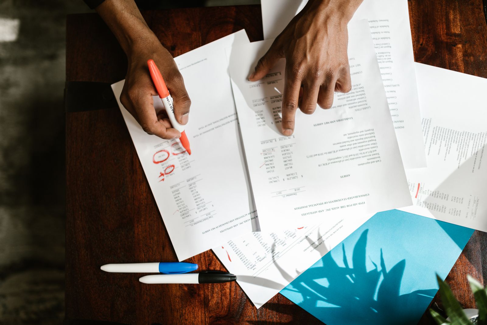 Overhead view of two hands going over printed financial statements on a dark wooden desk — one pointing at a column of figures, the other holding an orange marker beside numbers circled in red