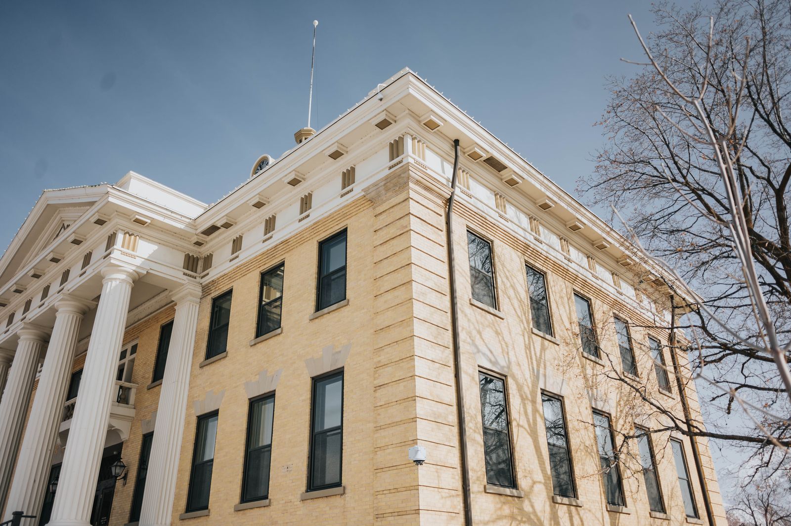 Low-angle view of the Box Elder County Courthouse in Brigham City, Utah — cream brick facade with white fluted columns and a dentiled cornice under a clear winter sky, bare tree branches at right
