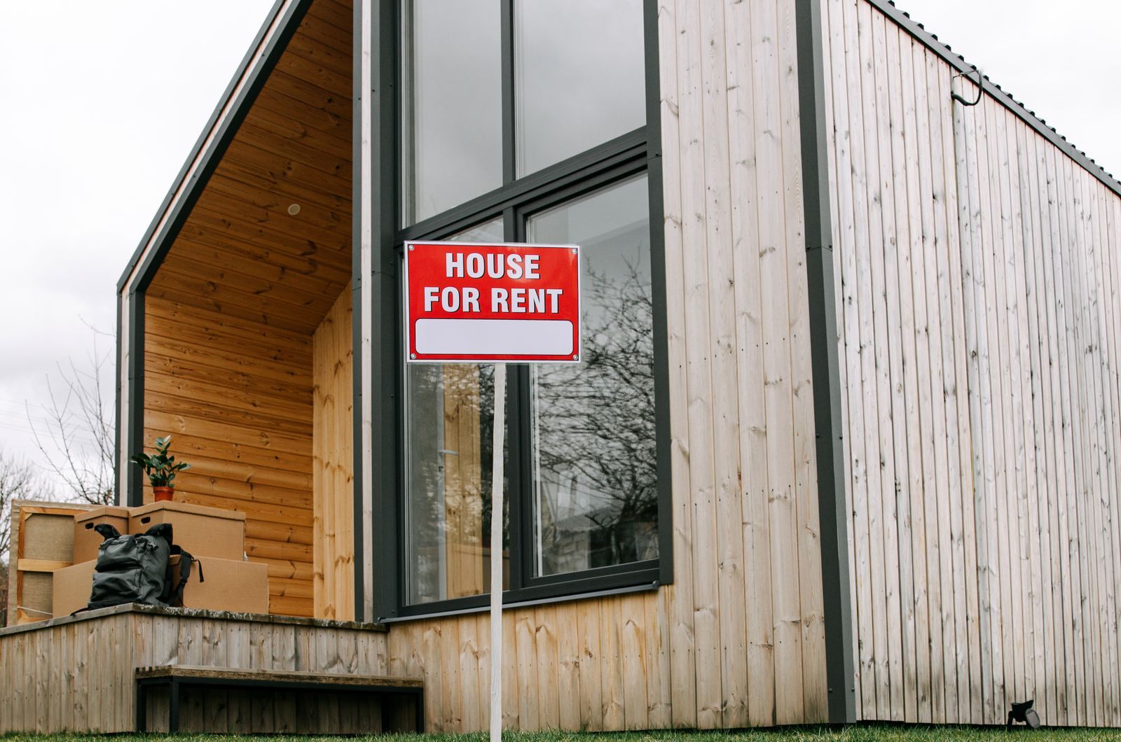 A red House for Rent sign staked in the lawn in front of a wood-sided house with tall black-framed windows and moving boxes stacked on the porch