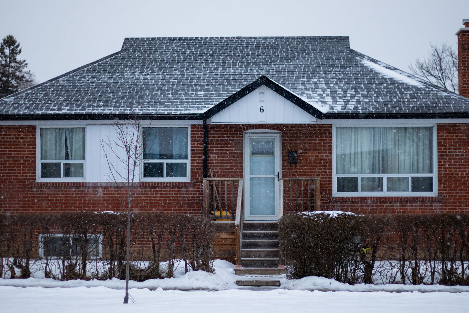 A residential brick home with snow falling and accumulated on the roof — the visual of a home that has been sitting on the market through winter