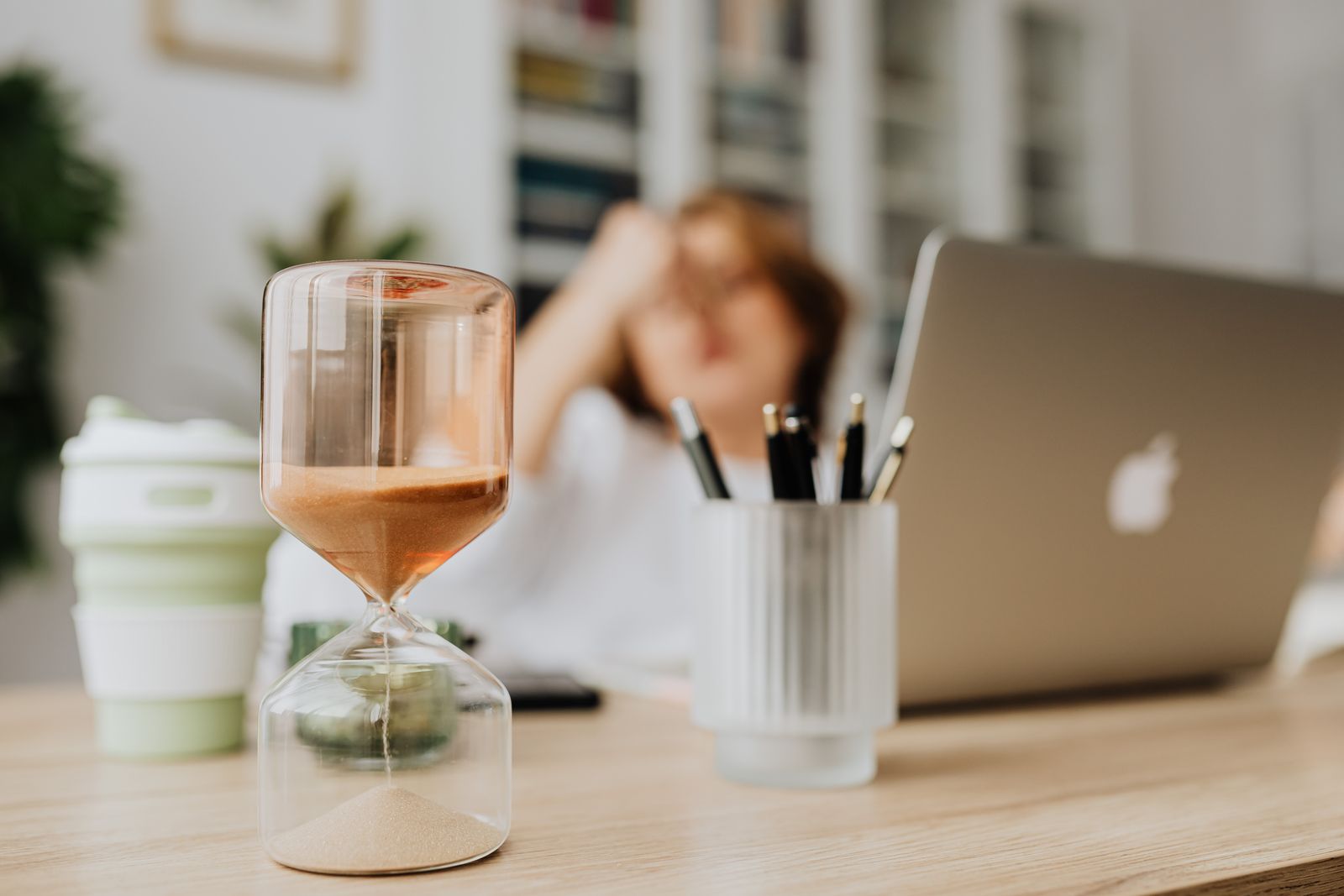 A glass hourglass with sand running through it sits in sharp focus on a light wood desk, with a person out of focus behind it resting their head on one hand beside a laptop