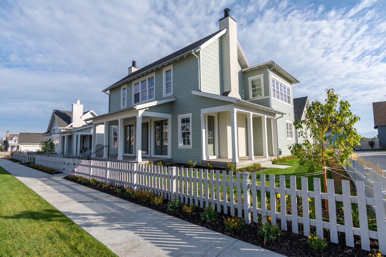 Two-story sage-green farmhouse with white trim, a deep covered front porch and a stucco chimney, fronted by a white picket fence and a concrete sidewalk in a newer master-planned neighborhood under a partly cloudy sky