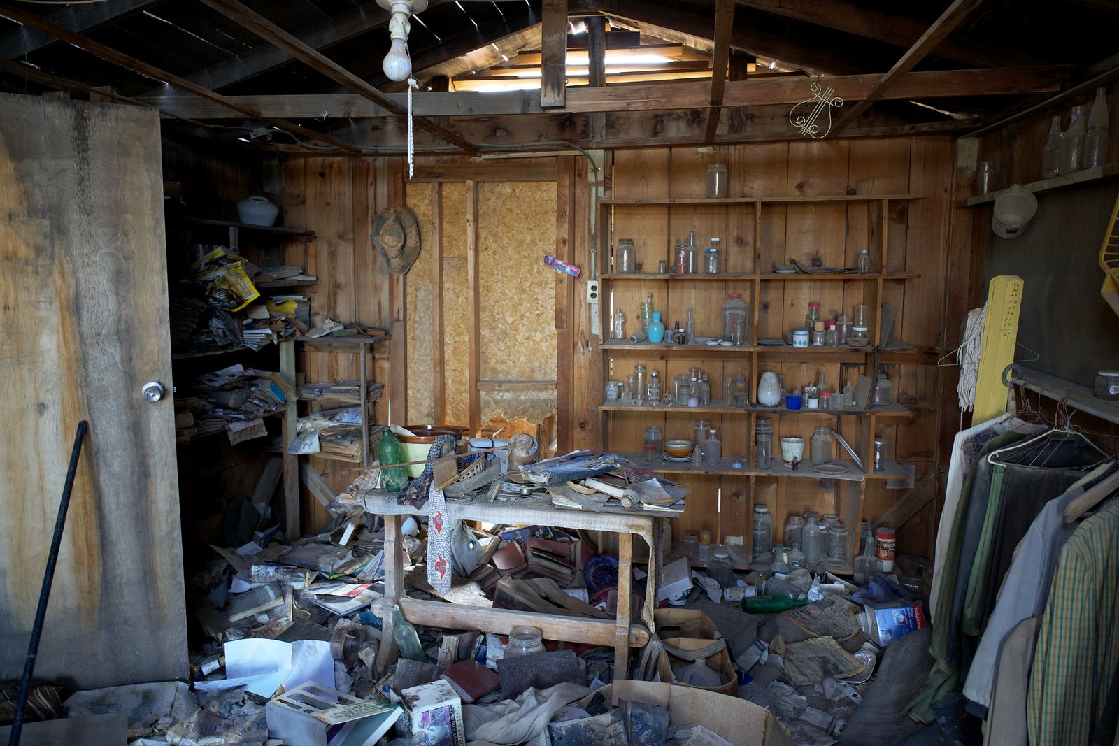 A wood-paneled room buried in debris — dusty jars on open shelves, a table piled with papers and bottles, clothes on a rack, and an exposed ceiling with a bare bulb