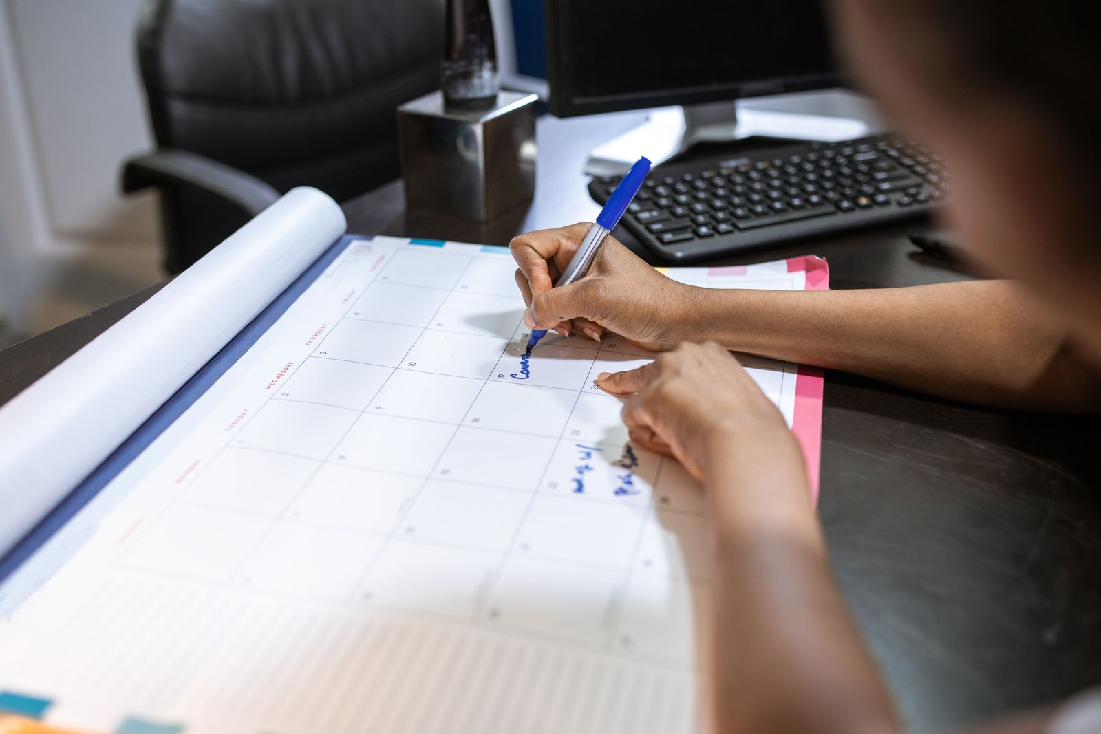 Someone writing appointments into the date squares of a large desk-pad month calendar with a blue pen, a keyboard and monitor on the desk behind it