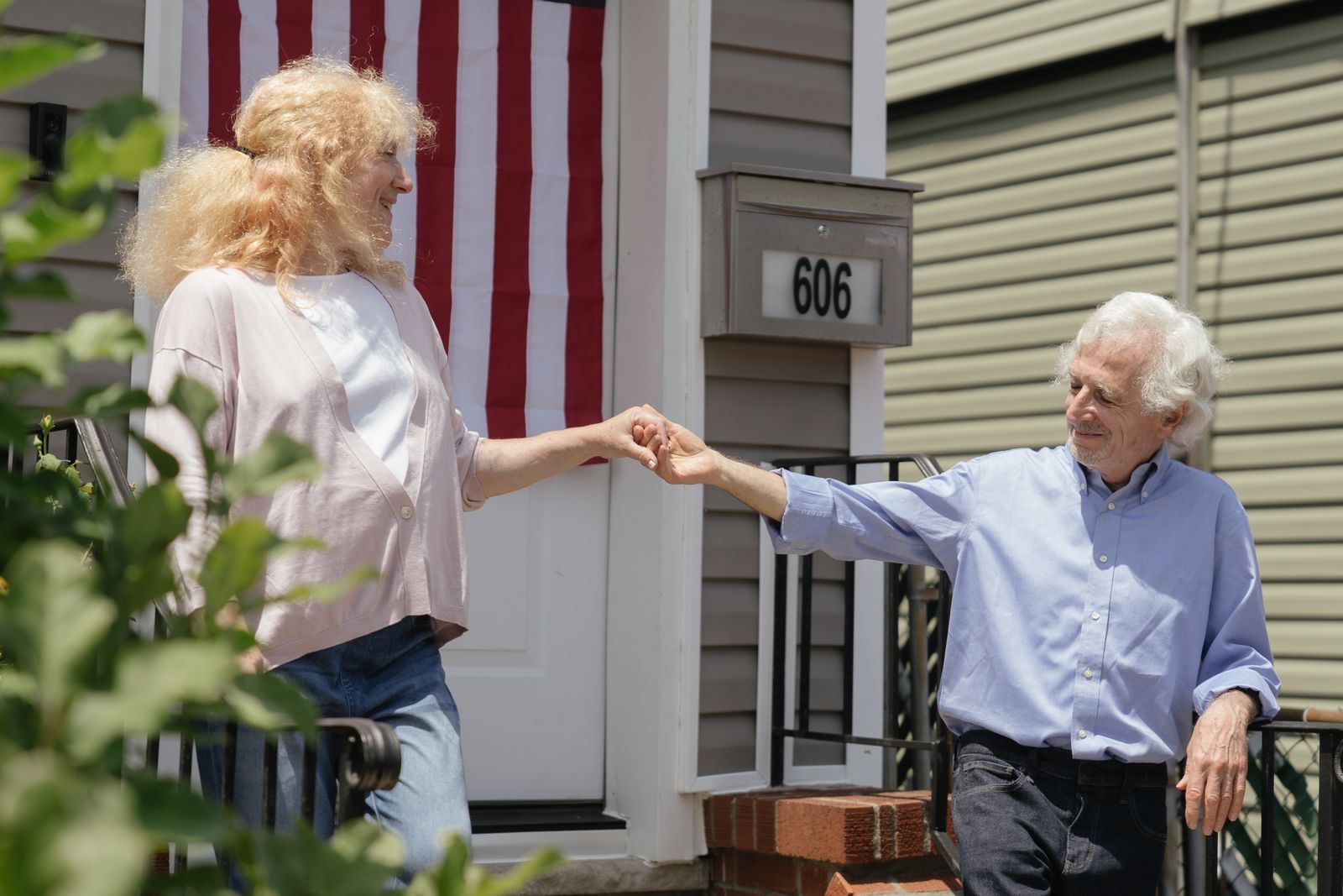 An older couple holding hands on the front steps of their sided house, a white front door and an American flag behind them