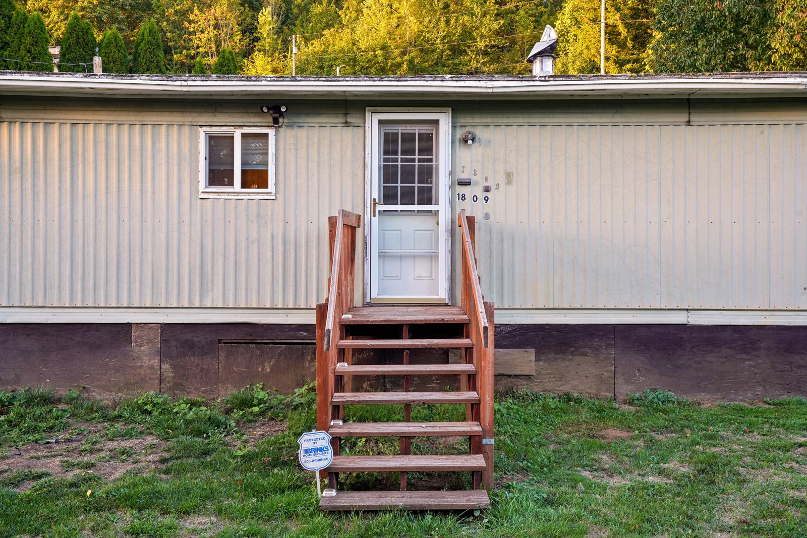 Exterior of a single-section manufactured home with light siding, skirting along the base, and a set of wooden entry steps leading to the front door on a sunny day