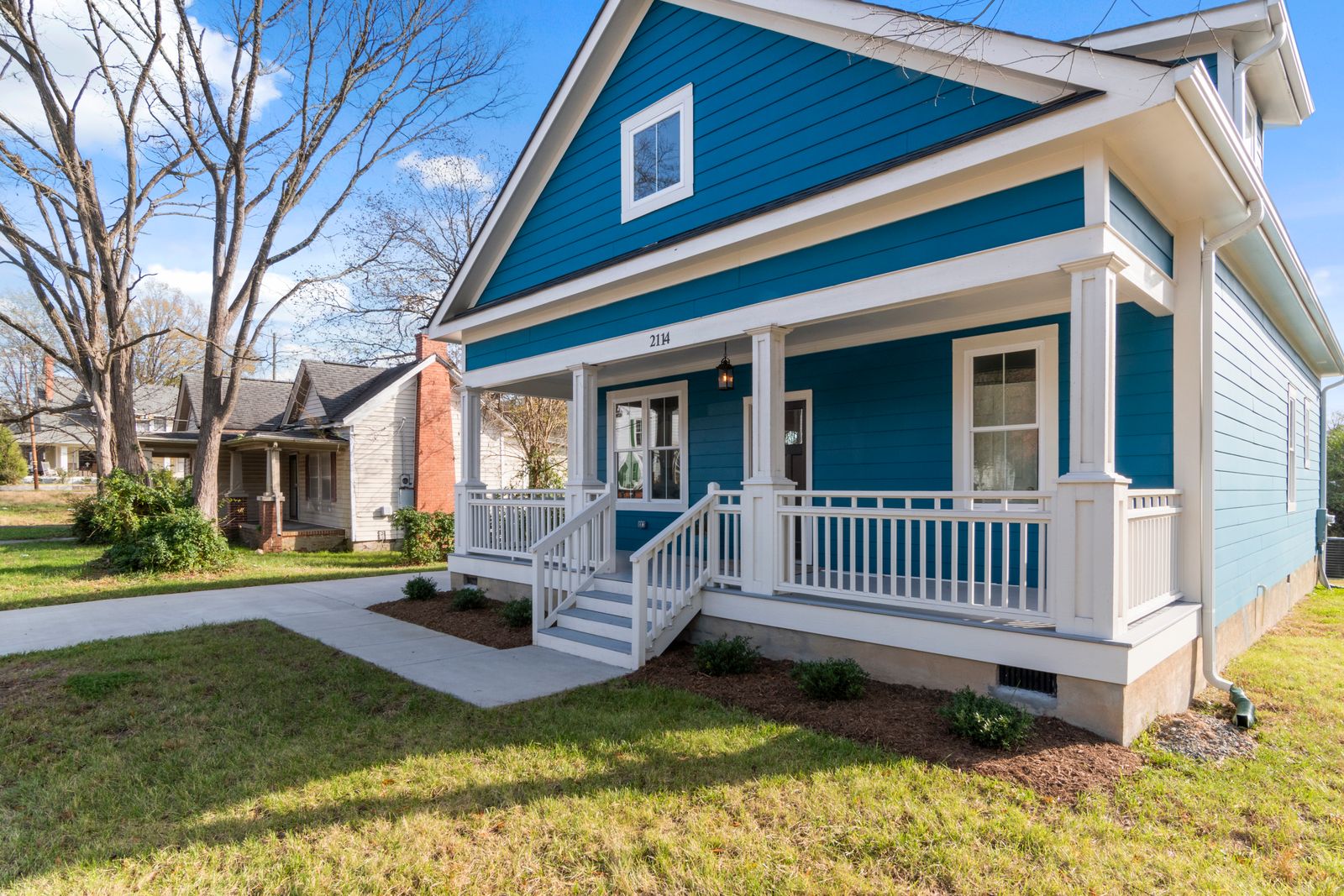 A teal blue bungalow with white trim and a covered front porch with white railings, house number 2114 above the porch, bare winter trees and a neighboring house behind it