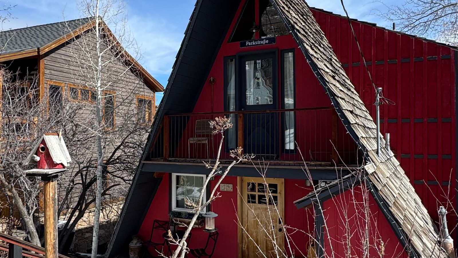 A red A-frame cabin with a steep pitched roof standing in deep snow among evergreen trees on a winter afternoon