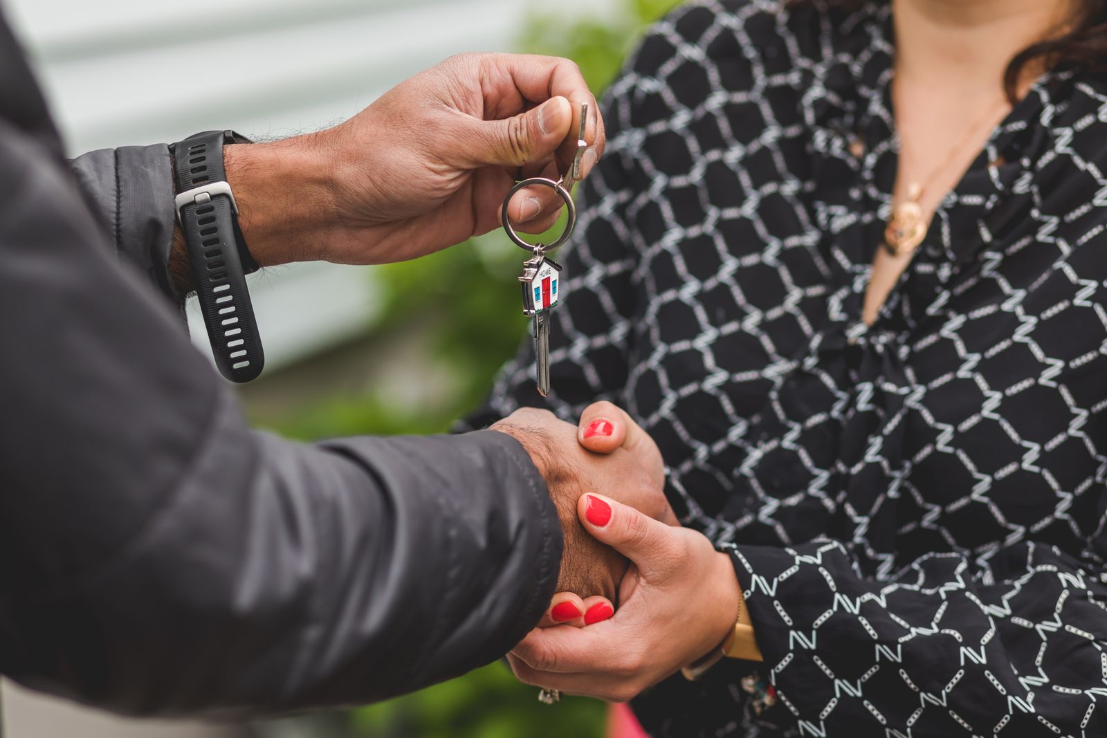 Close-up of one person handing a house key on a ring with a small house-shaped key cover to another, outdoors in front of a white-sided house